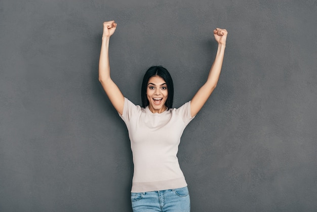 Photo she is a winner! happy young woman in casual wear keeping arms raised and looking at camera while standing against grey background