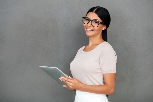 She knows the secret of success. Cheerful young businesswoman holding digital tablet and looking at camera while standing against grey background