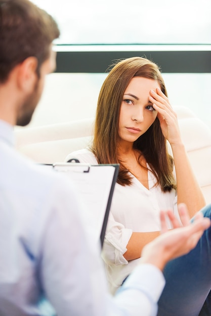 She needs to be consoled. Worried young woman sitting at the chair and holding hand on head while male psychiatrist sitting in front of her and gesturing