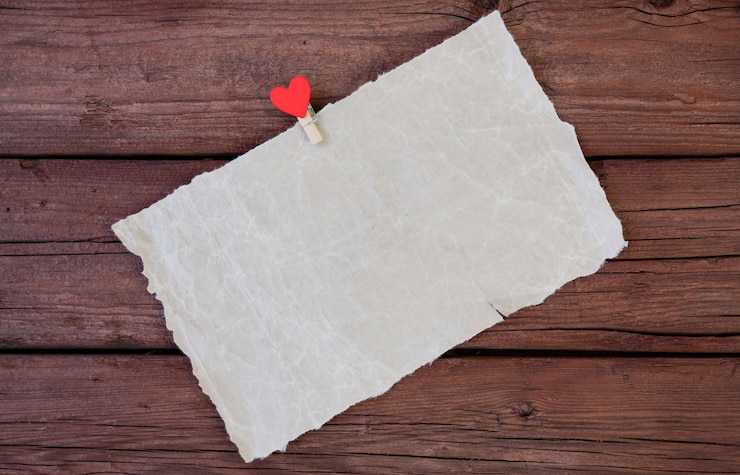 फोटो a sheet of parchment paper with a clothespin with a red heart on a wooden background. copy the space