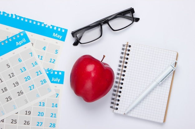 Листы ежемесячного календаря, красное яблоко, очки, блокнот на белом. Экономический расчет, калькуляция