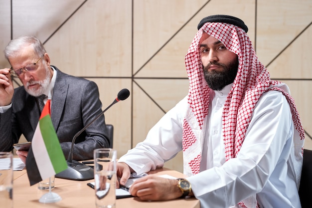 Sheikh man presenting his ideas to colleagues and listening for ideas for success investments at bright modern office room, sitting behind microphone and looking at camera