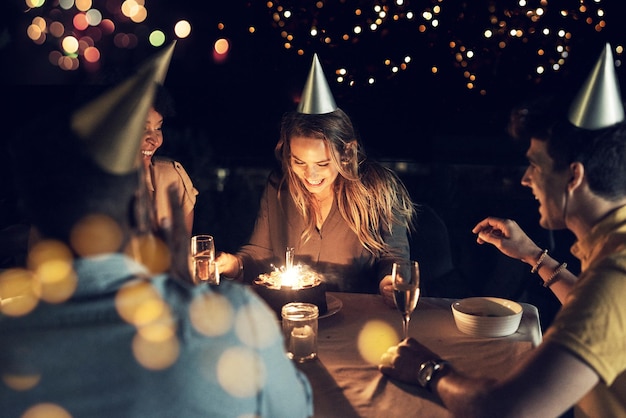 親しい友人全員と一緒に誕生日を祝う彼女は、夕方の屋外の集まりでテーブルの周りで一緒に誕生日を祝う友人のグループのショット