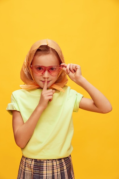 Shh beautiful girl child in stylish clothes headscarf and sunglasses posing over yellow studio