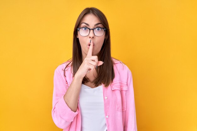 Shh brunette woman asks to keep quiet or secret keeps forefinger on lips isolated on yellow background