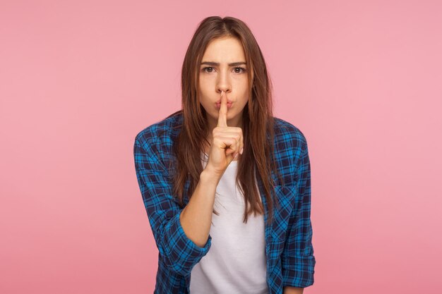 Shh don39t speak Portrait of worried unhappy girl in checkered shirt holding finger on lips showing silence gesture asking to stay quiet keep secret indoor studio shot isolated on pink background