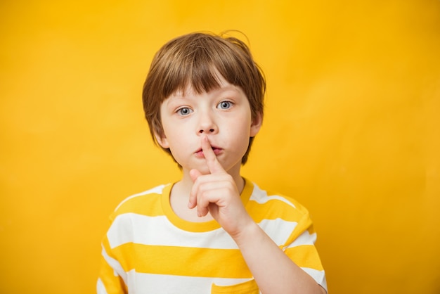 Shh, keep silence concept. Young caucasian child boy gesturing, hush sign posting standing isolated over yellow background. Studio shot. Finger on lips, silent gesture, secret. Copy space