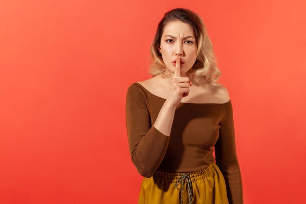 Shh, shut up. Serious and angry woman with blonde hair in brown casual blouse showing gesture secret sign with finger near her lips. Expression emotion. Indoor isolated on red background