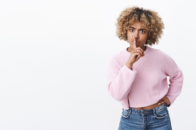 Shh you cannot swear here. Angry and serious-looking strict elder sister with afro haircut frowning shushing with index finger over folded lips demanding silence and keep secret over white wall