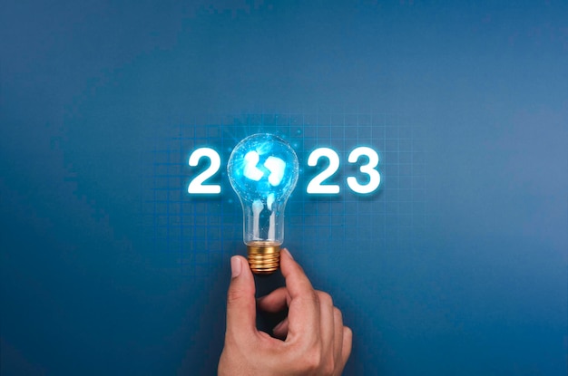 Shining 2023 calendar year numbers neon style with creative trend light bulb holding by businessman's hand on digital network and blue background Happy 2022 New Year greeting card with light bulb