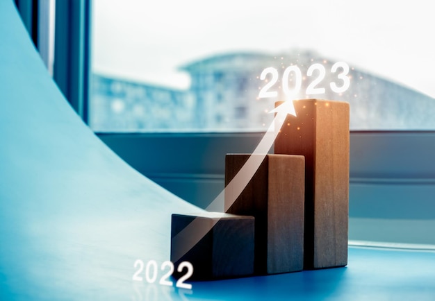 Shining rise up arrow on wood blocks chart steps as a graph from year 2022 to 2023 on blue background with cityscape business growth process profit wealth trends economic improvement concepts