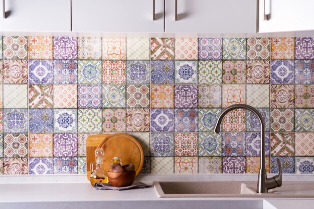 Shiny stainless steel faucet with chrome water tap, stone sink and wooden bowls with olive olives on a wall of colored old tiles. Kitchen interior