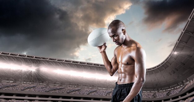 Shirtless african american male rugby player holding a rugby ball against stadium in background