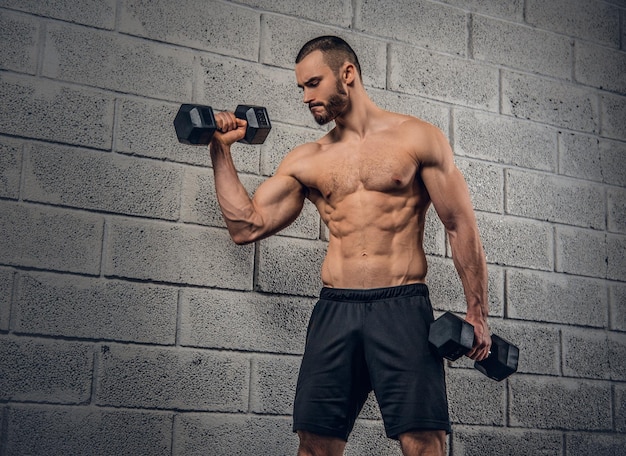 Фото Спортивный бородатый мужчина без рубашки тренируется с гантелями на фоне серой стены.