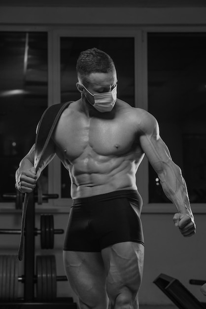 A shirtless bodybuilder in a medical face mask is posing after\
a workout in gym