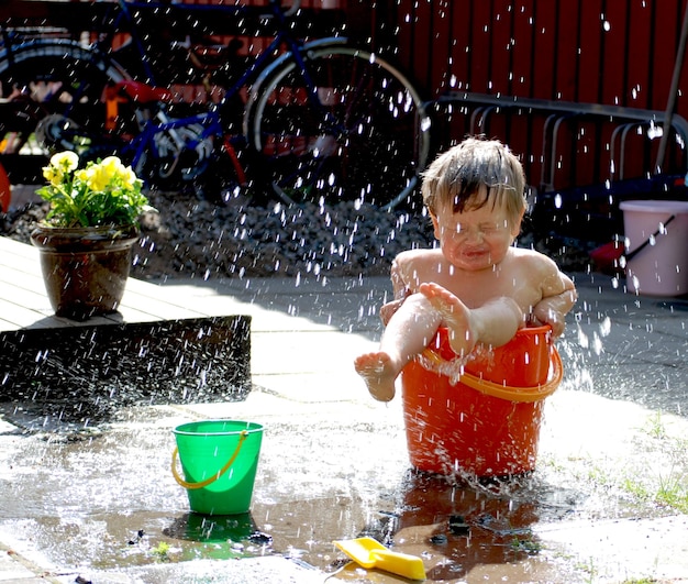 シャツを着ていない少年が庭のバケツに水をこぼしている