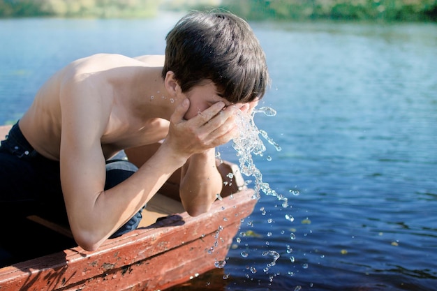 ボートで航海している間シャツなしの少年が湖から顔を洗っている