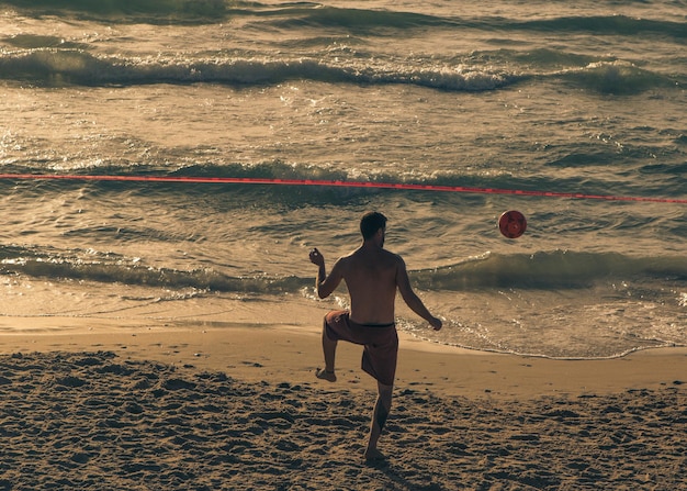 Foto uomo senza camicia sulla spiaggia che gioca con una palla le onde spazzano la sabbia ancora calda