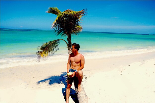 Shirtless man holding coconut while sitting on palm tree at beach