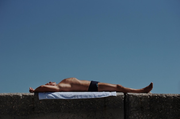 shirtless man relaxing on retaining wall against clear blue sky