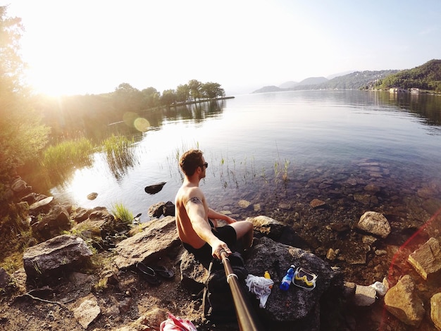 Photo shirtless man taking selfie using monopod while sitting on rock by lake