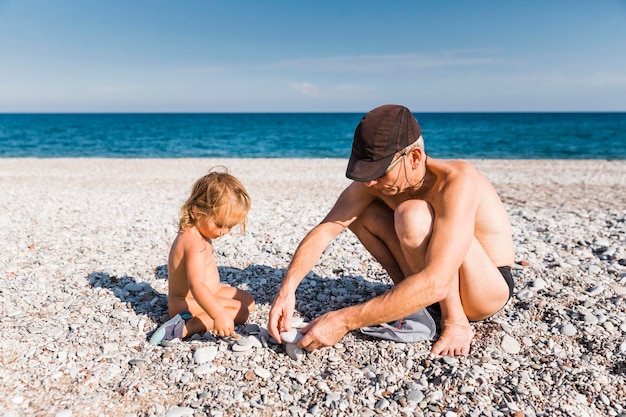 Photo shirtless man with grandchild crouching at beach on sunny day