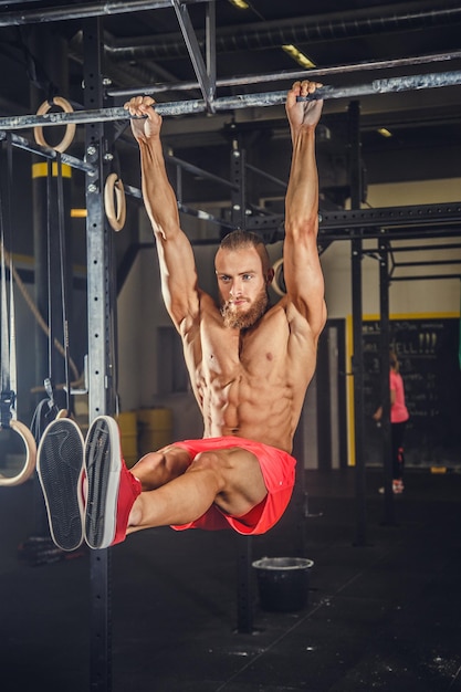 Shirtless muscular bearded male doing exercises on horizontal bar in a gym club.