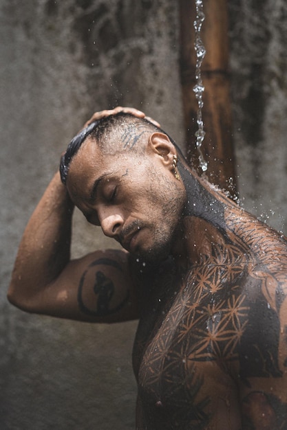 문신이 있는 벗은 근육질의 라틴 아메리카 남성은 열대 나뭇잎 배경에서 운동한 후 샤워를 하고 있습니다. 전용 빌라에 샤워 시설이 있습니다.