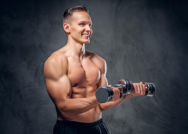 The shirtless muscular male holds a set of dumbbells over dark grey vignette background.