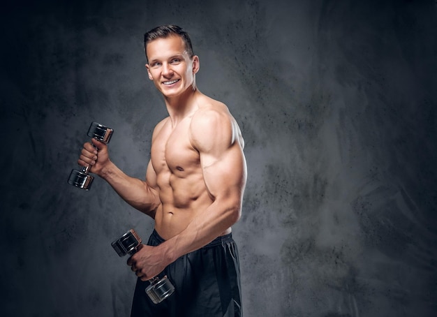 The shirtless muscular male holds a set of dumbbells over dark grey vignette background.