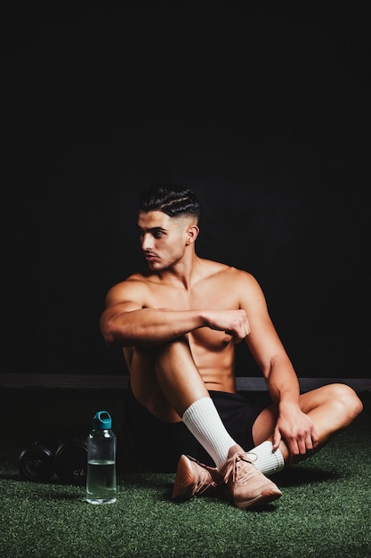 Shirtless muscular man sitting on the floor of the gym next to some weights