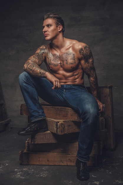 Shirtless muscular man with tattooes on his body sitting on wooden boxes.