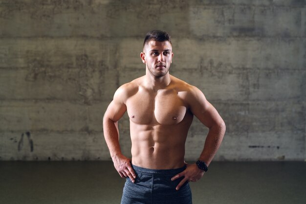 Photo shirtless serious caucasian man with hands on hips posing while standing in front of the wall.