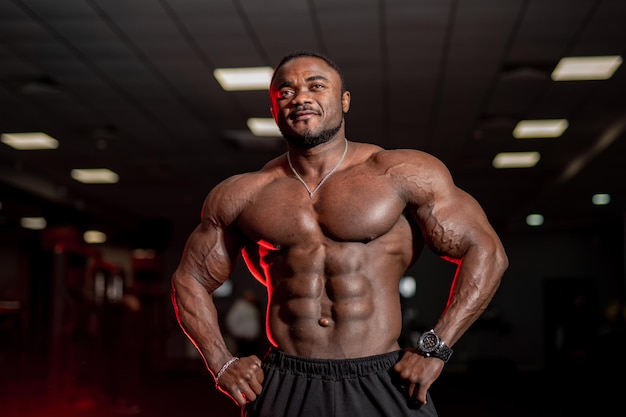 Shirtless strong bodybuilder with perfect abs poses to the camera. Modern gym background. African american athlete.