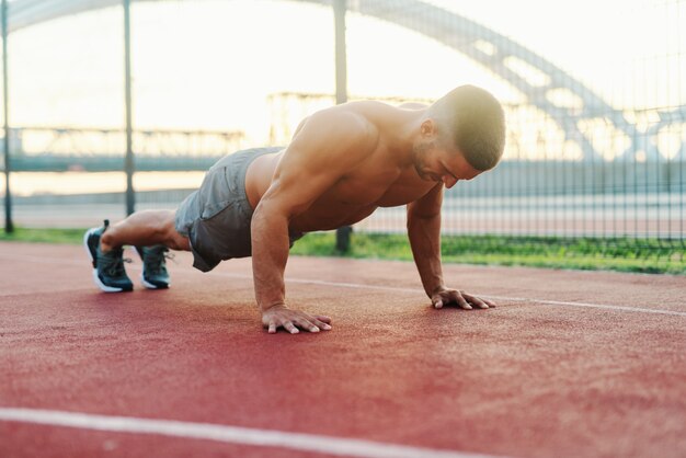 Shirtless strong dedicated muscular man doing push ups on the court in the morning.