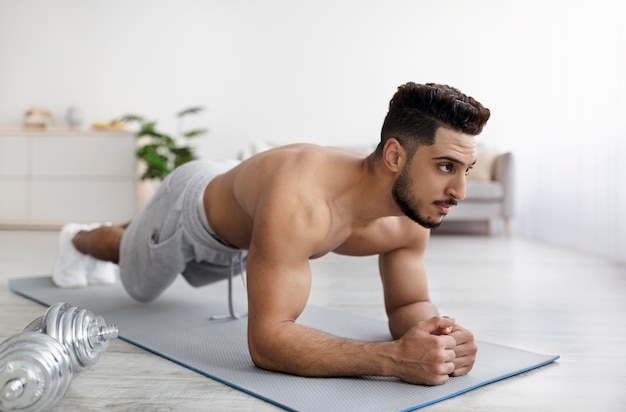 Shirtless young arab man standing in elbow plank on yoga mat exercising abs muscles on domestic