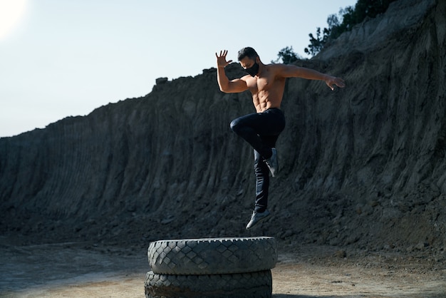 Фото Молодой культурист без рубашки в черной маске использует большое тяжелое колесо для вечерней тренировки. сильный человек прыгает высоко во время тренировки среди песочницы.