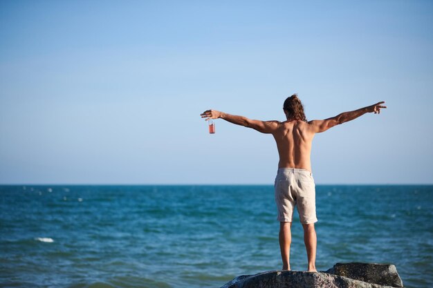 Giovane senza camicia in piedi sulla spiaggia rocciosa con in mano una bottiglia di bibita analcolica che allunga le braccia e lo sguardo