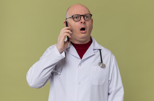 Uomo adulto scioccato con gli occhiali in uniforme da medico con stetoscopio che parla al telefono