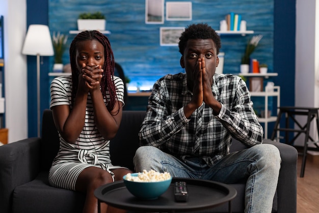 Shocked african american couple watching tragic news on television program while sitting at home. Terrified people reacting bad while watching horror movies sitting alone on couch.