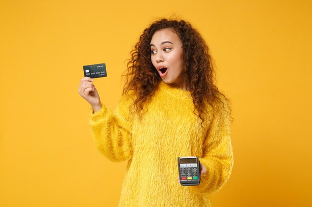 Shocked african american girl posing isolated on yellow background. People lifestyle concept. Mock up copy space. Holding wireless modern bank payment terminal to process acquire credit card payments.