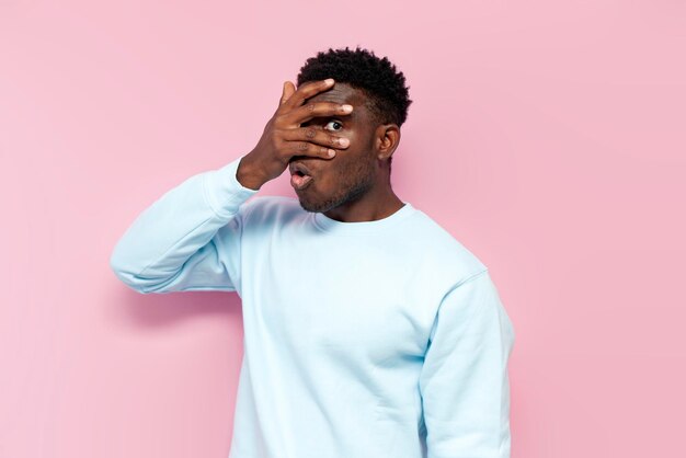 shocked african american man in blue sweater covers his face with his hands hiding and peeking