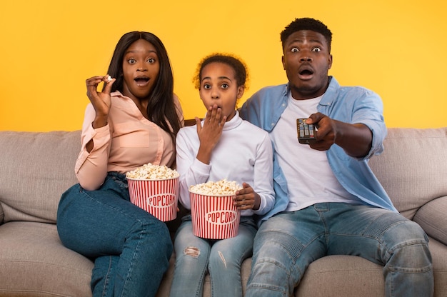 Shocked african american parents and their daughter eating\
popcorn and watching movie on tv sitting on couch