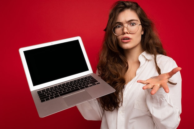 Scioccato stupito withless bella bruna riccia giovane donna sayong wow che tiene computer portatile con gli occhiali camicia bianca che guarda l'obbiettivo isolato sopra priorità bassa rossa della parete. modello