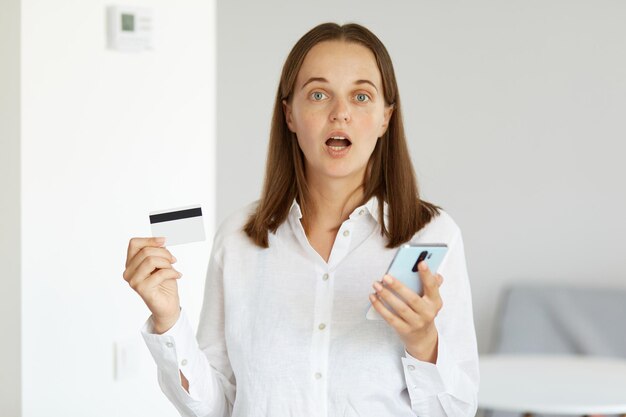 Shocked amazed woman wearing white shirt standing indoor in light room holding credit card and cell phone in hands, being surprised of winning lottery or betting.