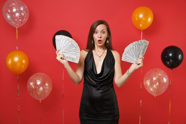 Shocked amazed young woman in little black dress celebrating, holding bundle lots of dollars, cash money on bright red background air balloons. Happy New Year, birthday mockup holiday party concept.