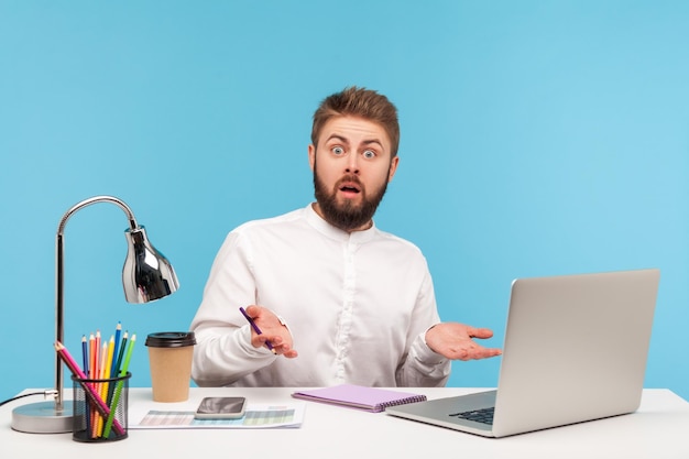 Scioccato infastidito impiegato uomo che diffonde le mani guardando la fotocamera con stupore seduto sul posto di lavoro con laptop e caffè lavoro stressante studio indoor girato isolato su sfondo blu