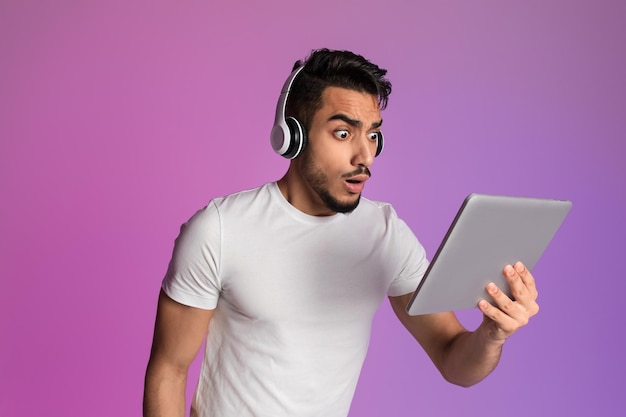 Shocked arab guy in headphones looking at tablet screen cannot believe his eyes winning online