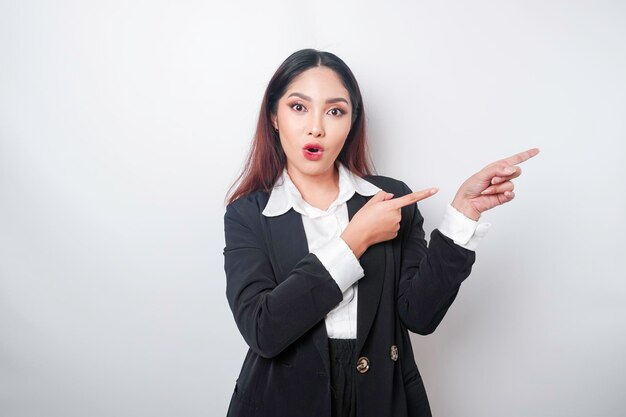 白い背景で隔離された彼女の横にあるコピー スペースを指して黒のスーツを着てショックを受けたアジアの実業家