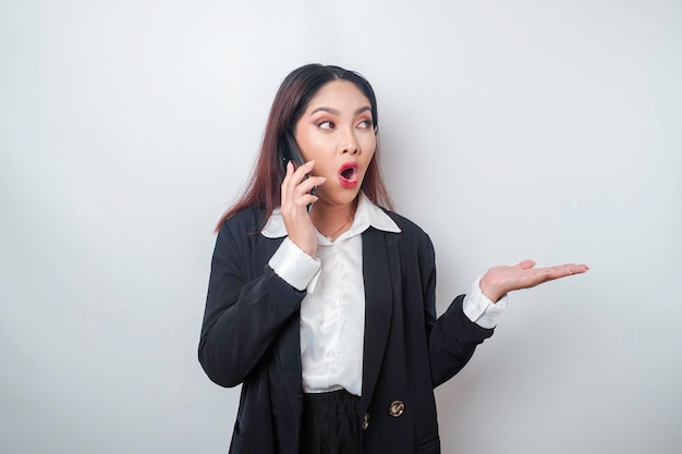 Shocked Asian businesswoman wearing a black suit pointing at the copy space beside her while talking on her phone isolated by a white background